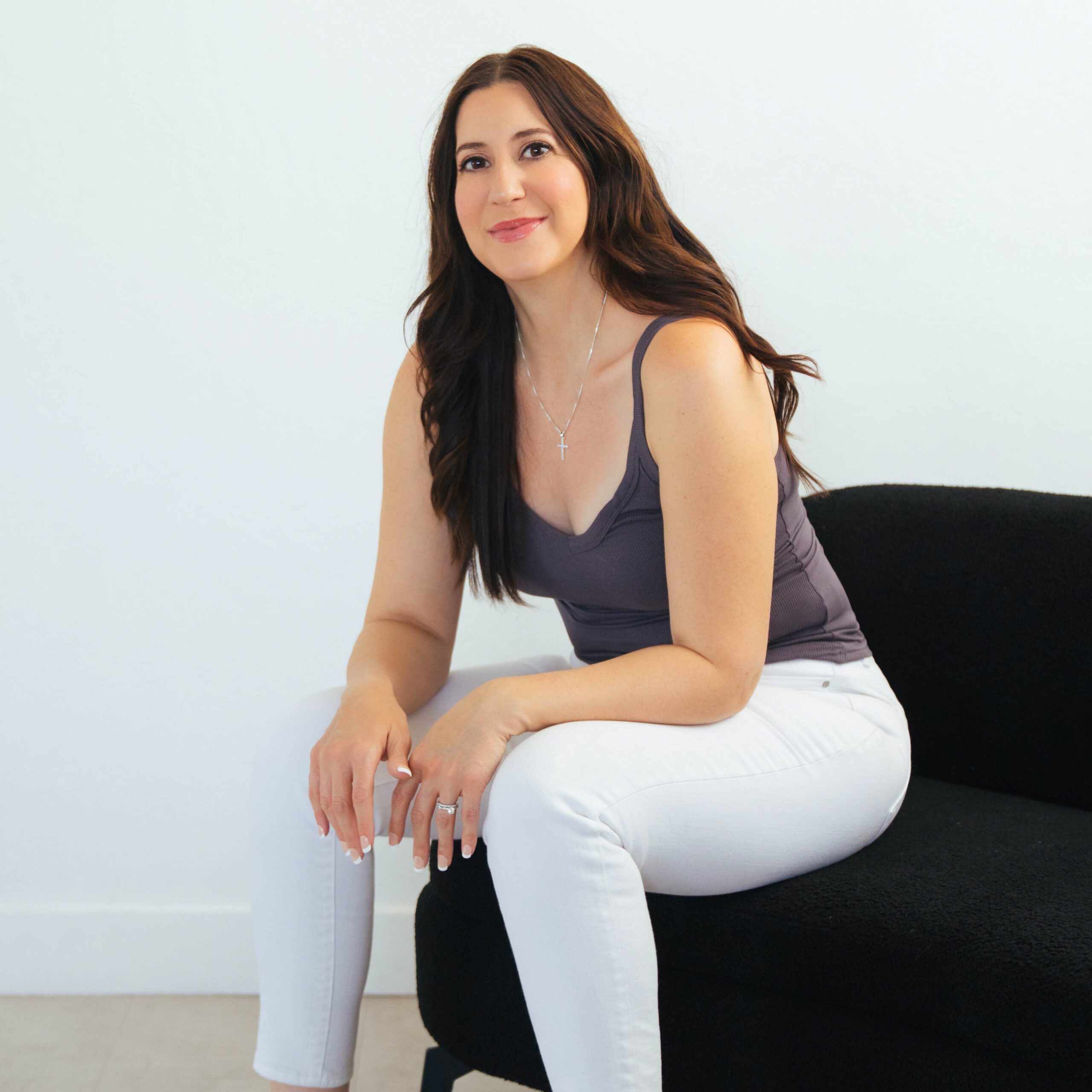 Woman with long dark hair sitting forward on a chair with her arms resting on her legs. She's wearing a blue tank top and white leggings.
